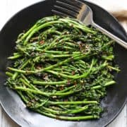 Grilled broccolini is served on a dark plate with a fork.