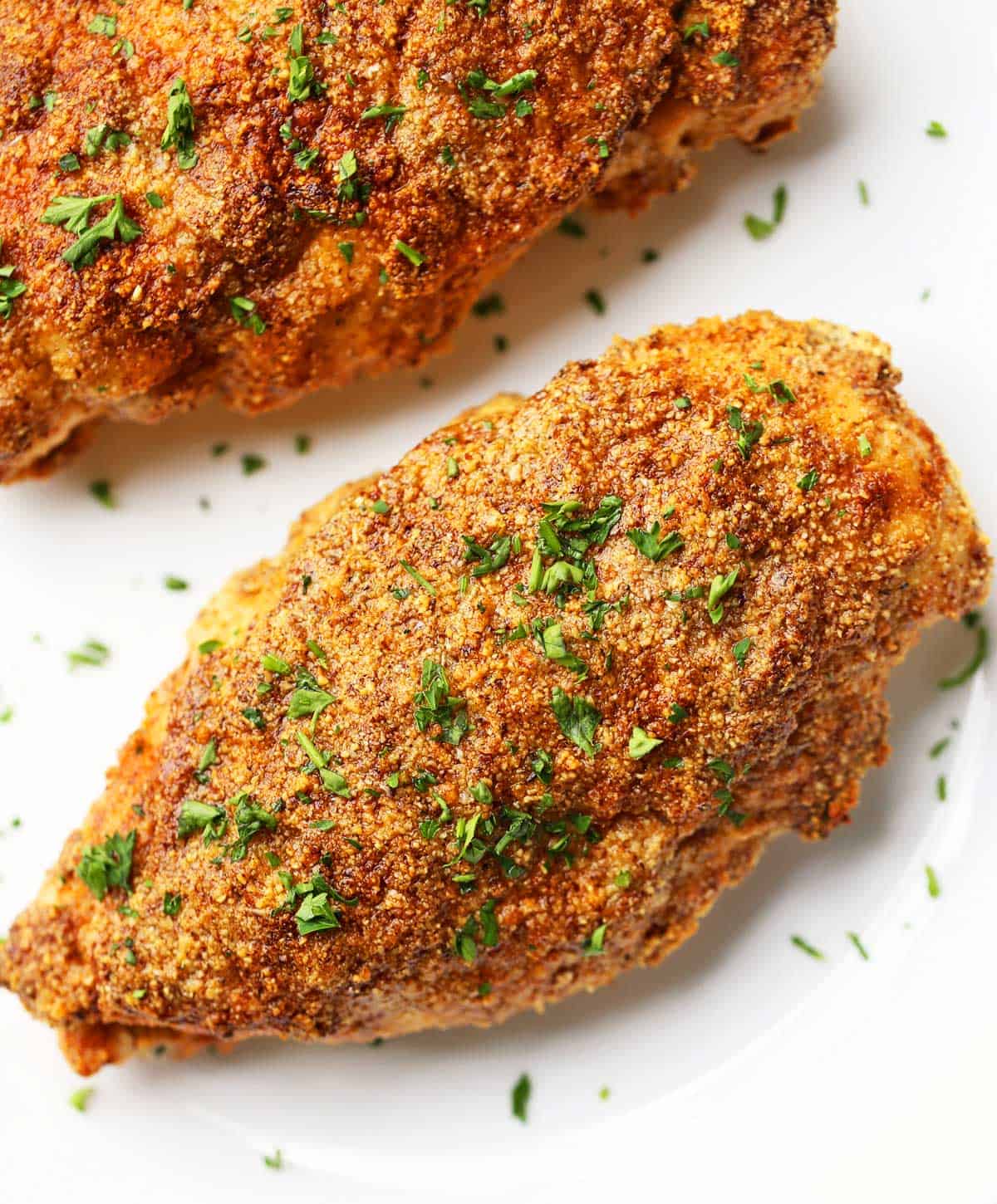 Two pieces of parmesan-crusted chicken are topped with parsley and served on a white plate. 