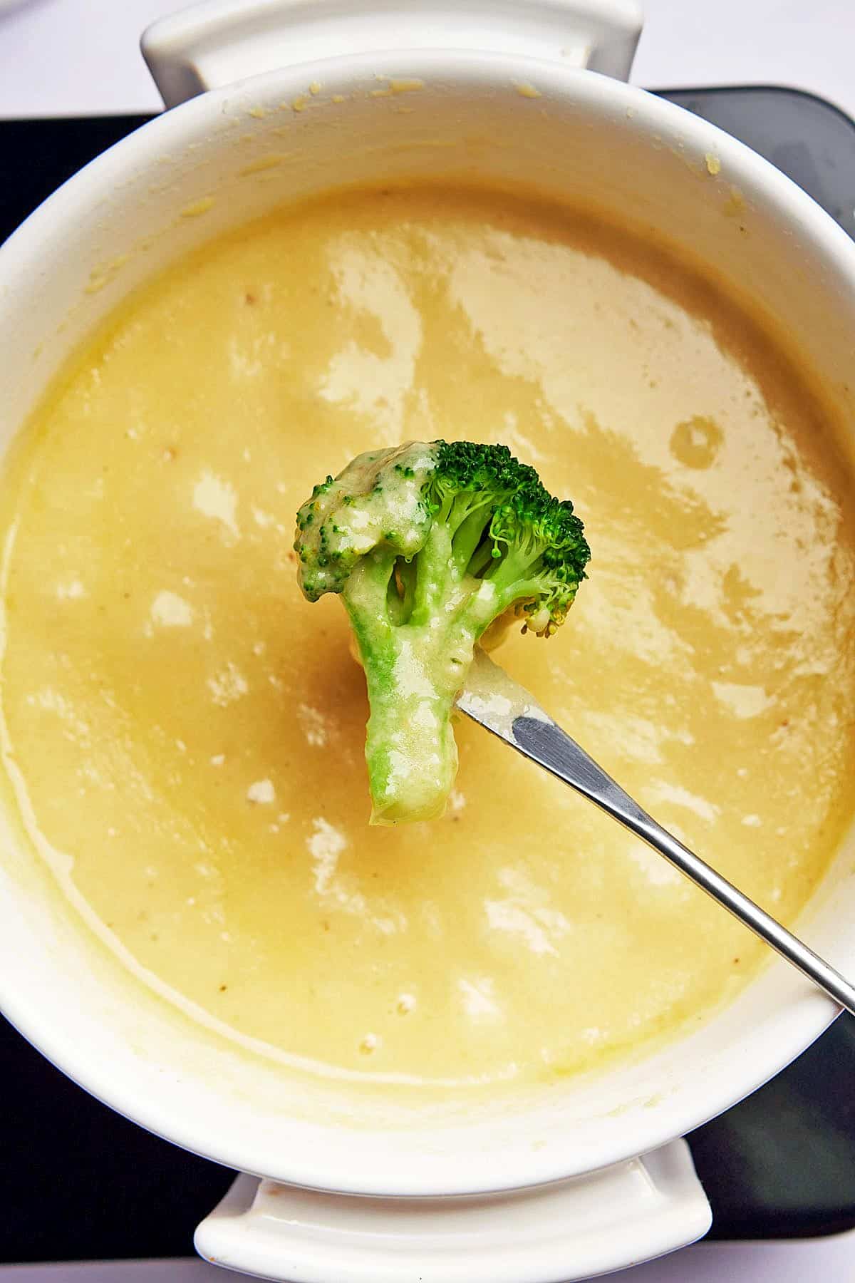 Closeup of a broccoli floret dipped into a cheese fondue. 