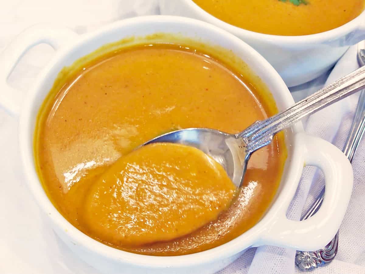 Pumpkin curry soup is served in a white bowl. A spoon scoops it out to show its creaminess. 